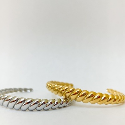 Duas pulseiras metálicas torcidas, uma prateada e outra dourada, sobre fundo branco