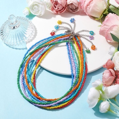 Colares coloridos de contas pequenas com fecho metálico e pingentes de flores sobre prato branco e fundo azul com flores artificiais