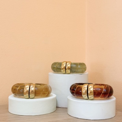 Pulseiras em resina verde, castanho e âmbar com detalhes dourados, expostas em suportes brancos redondos, fundo cor pêssego.