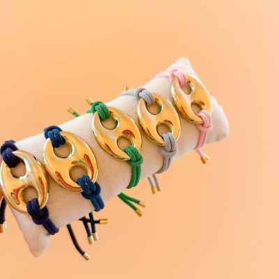 Pulseiras douradas com cordões coloridos exibidas numa almofada clara sobre fundo laranja