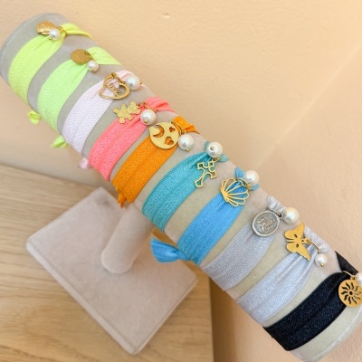 Pulseiras elásticas coloridas com pendentes dourados e pérolas num suporte creme.