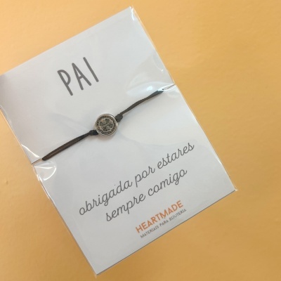 Pulseira preta com medalha prateada em cartão branco com texto para pai