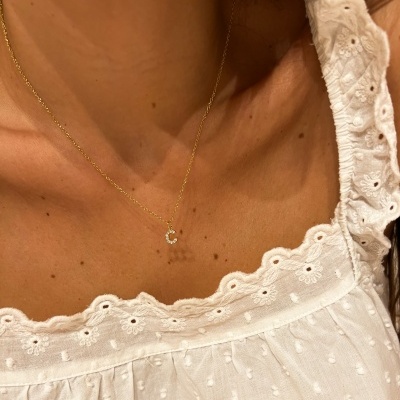 Colar dourado com letra 'C' em pedras brilhantes sobre blusa branca bordada