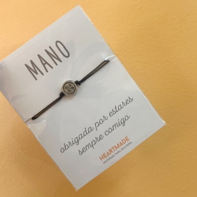 Pulseira com cordão preto e medalha metálica circular em embalagem branca com texto