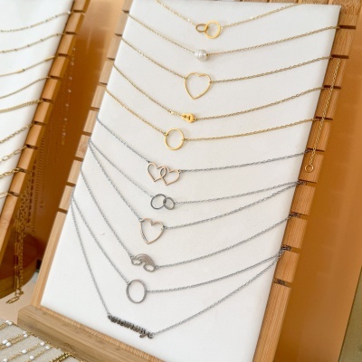 Colares finos em dourado e prata com formas decorativas exibidos em expositor branco e estrutura de madeira.