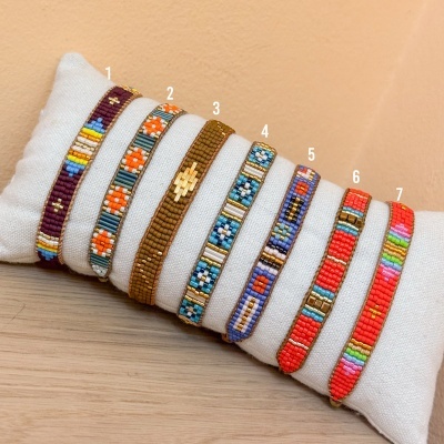Sete pulseiras coloridas de contas dispostas em almofada branca sobre superfície de madeira.