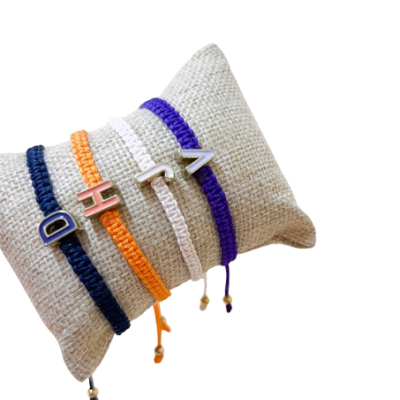 Pulseiras trançadas coloridas com letras metálicas sobre almofada bege