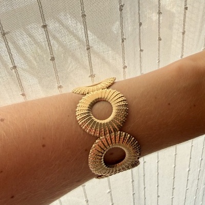 Pulseira dourada texturizada em pulso com fundo de cortina bege translúcida