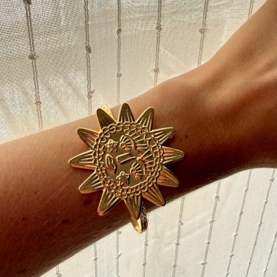 Pulseira dourada formato sol com gravações no pulso com cortina branca ao fundo