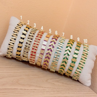 Dez pulseiras douradas com detalhes coloridos apresentadas em almofada branca.