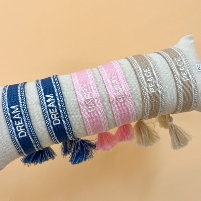 Pulseiras de tecido azul, rosa e bege com palavras bordadas e franjas, sobre almofada bege.