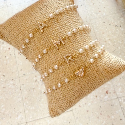 Três pulseiras de pérolas com letras douradas e um coração em almofada de tecido rústico.