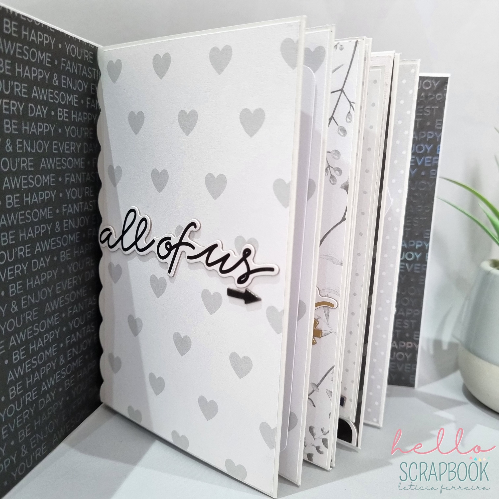 Álbum de scrapbook branco com corações cinza e texto 'all of us' na capa, em fundo com textos em inglês