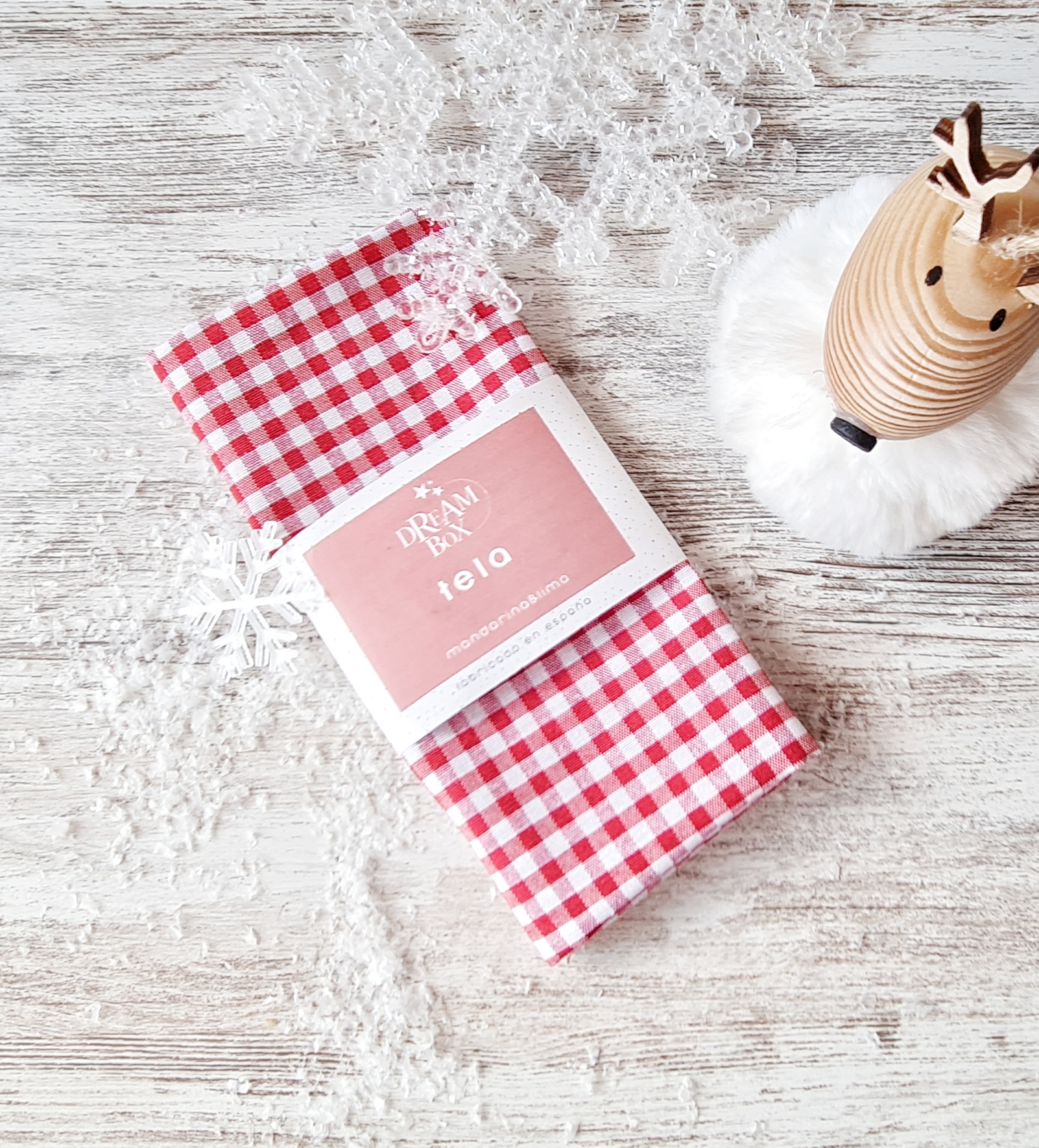 Pano de cozinha xadrez vermelho e branco com etiqueta rosa, ao lado de figura decorativa de rena em madeira