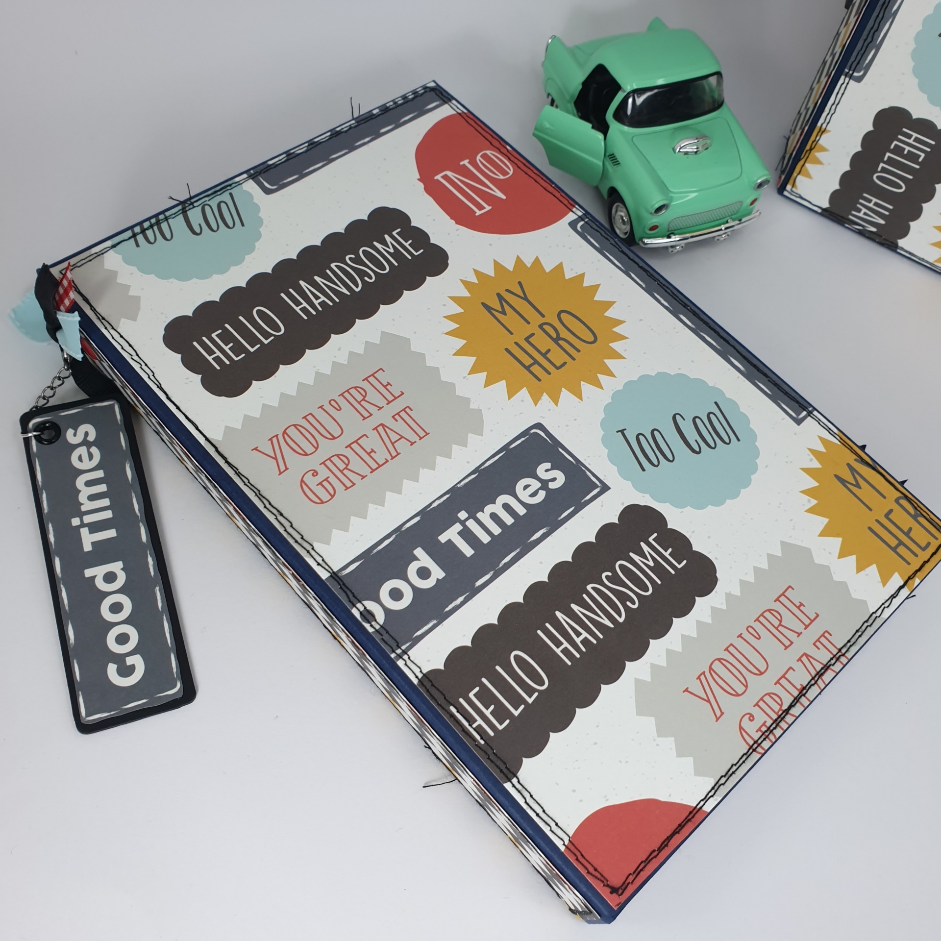 Livro ou caderno colorido com frases em inglês e chaveiro 'Good Times' ao lado de brinquedo de carro verde