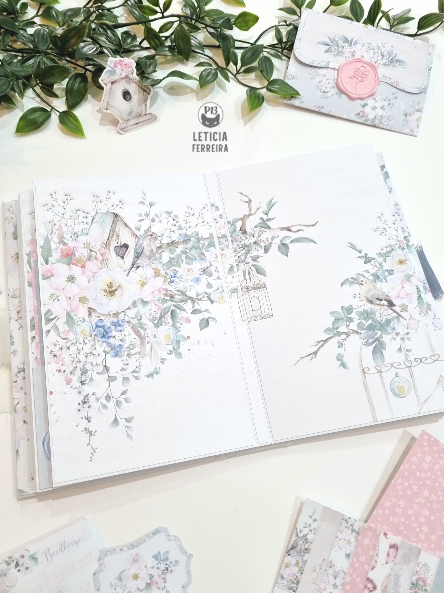 Caderno ilustrado aberto com flores e pássaros, envelope decorativo e plantas sobre mesa branca