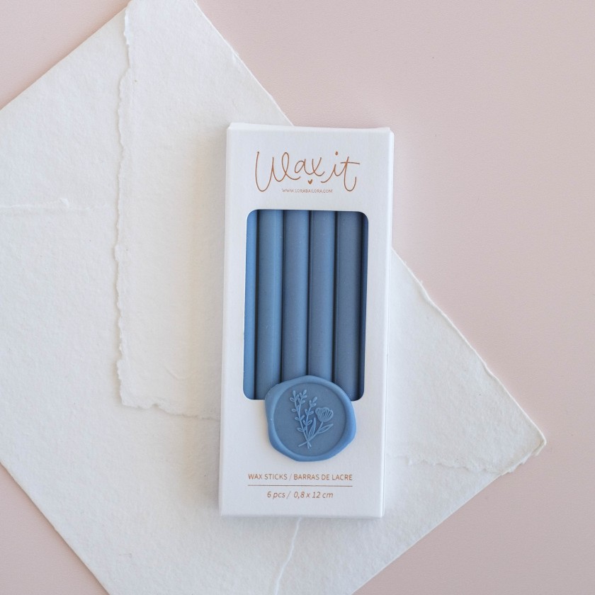 Caixa branca com 6 varetas de cera azul e selo de cera azul, sobre papel branco e fundo rosa.