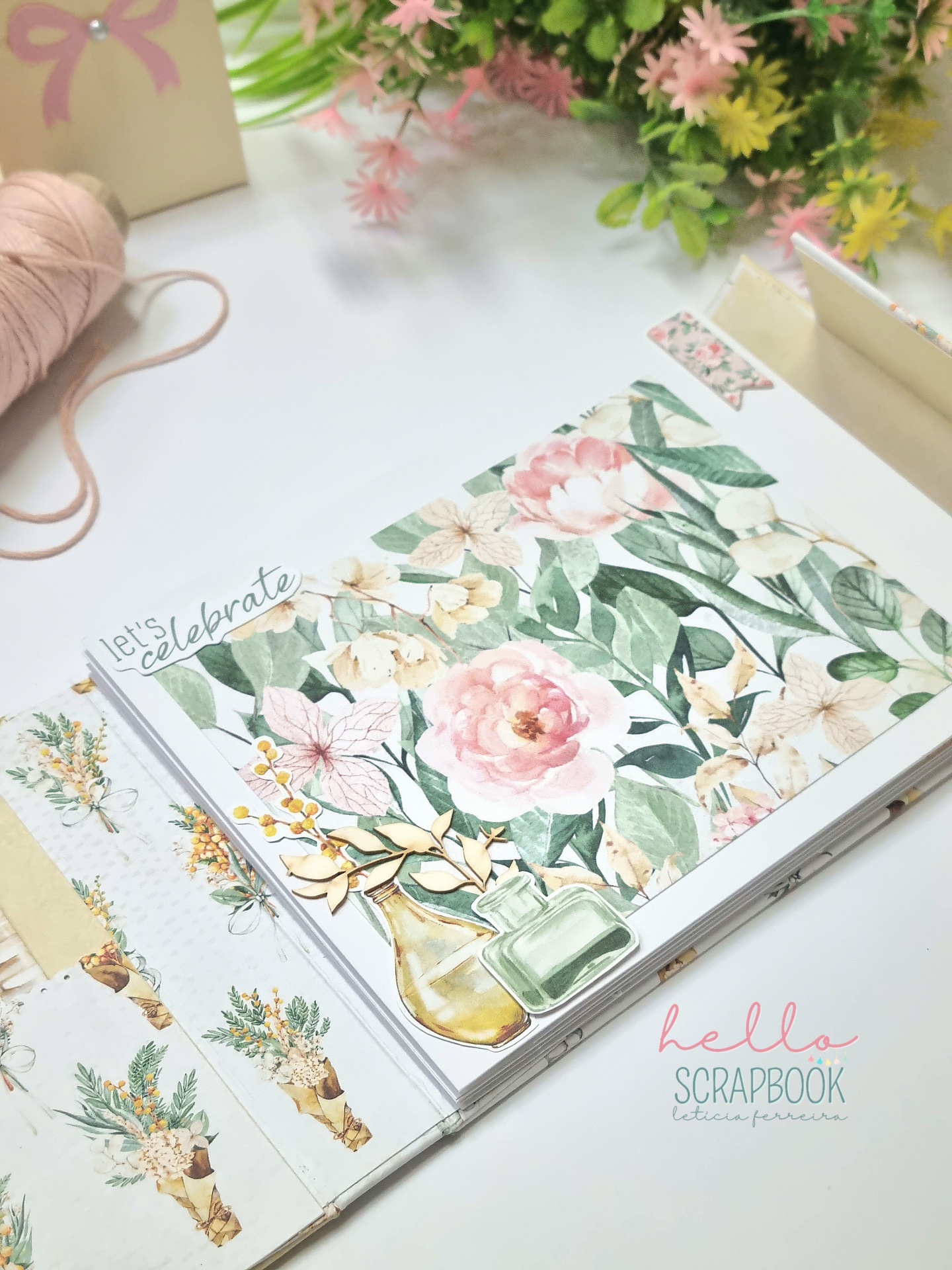 Caderno scrapbook com flores rosa e verdes, texto 'Let's Celebrate' e 'hello SCRAPBOOK', flores artificiais e fio rosa ao redor