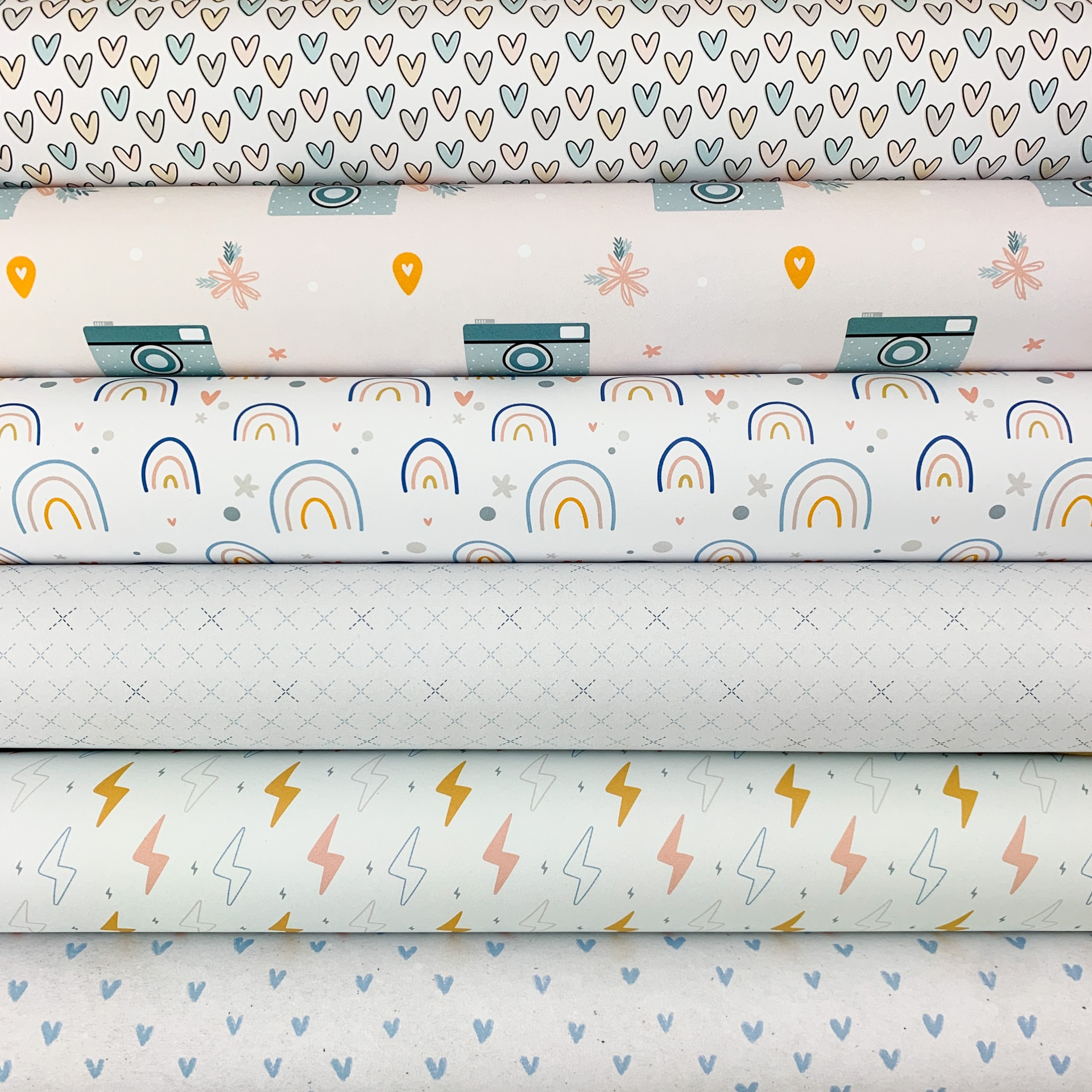 Tecidos com padrões variados de corações, câmeras, arcos-íris, pequenas cruzes e relâmpagos em tons de cinza, azul, laranja e terracota.