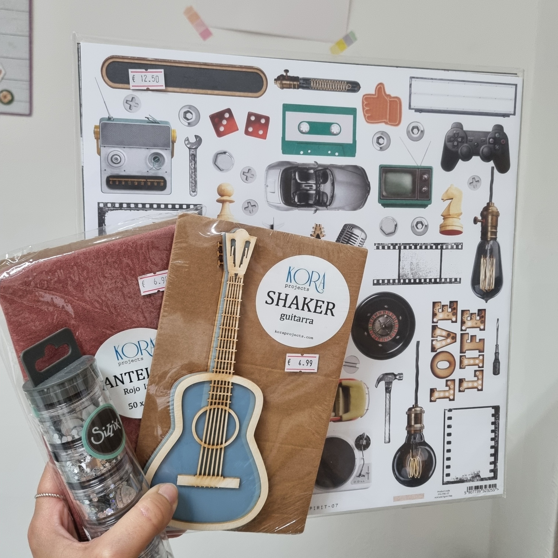 Mãos segurando botões de metal Sizzix e cartão com guitarra KORA projects SHAKER guitarra, com poster de objetos retro ao fundo.