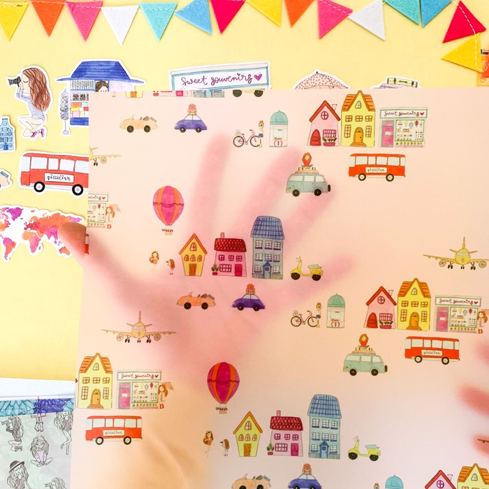 folha decorativa com desenhos de casas, veículos e balões, sombra de mão por trás, mesa amarela com bandeirolas