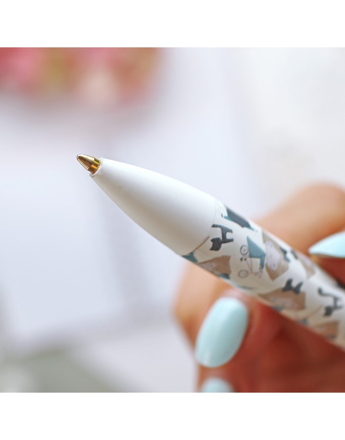Caneta esferográfica branca decorada com padrão de cães e ponteira dourada, segurada por mão com unhas azuis.