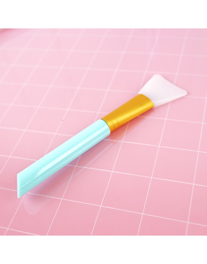 Espátula de silicone azul claro, dourada e branca sobre fundo rosa quadriculado