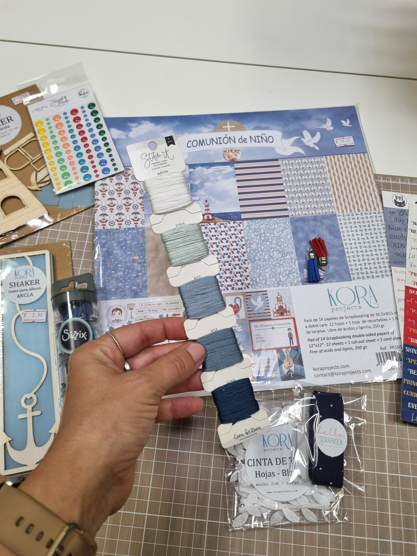 Materiais para scrapbook com papéis temáticos de comunhão infantil, fios de bordado azuis, adesivos coloridos e acessórios de artesanato sobre superfície branca e quadriculada.
