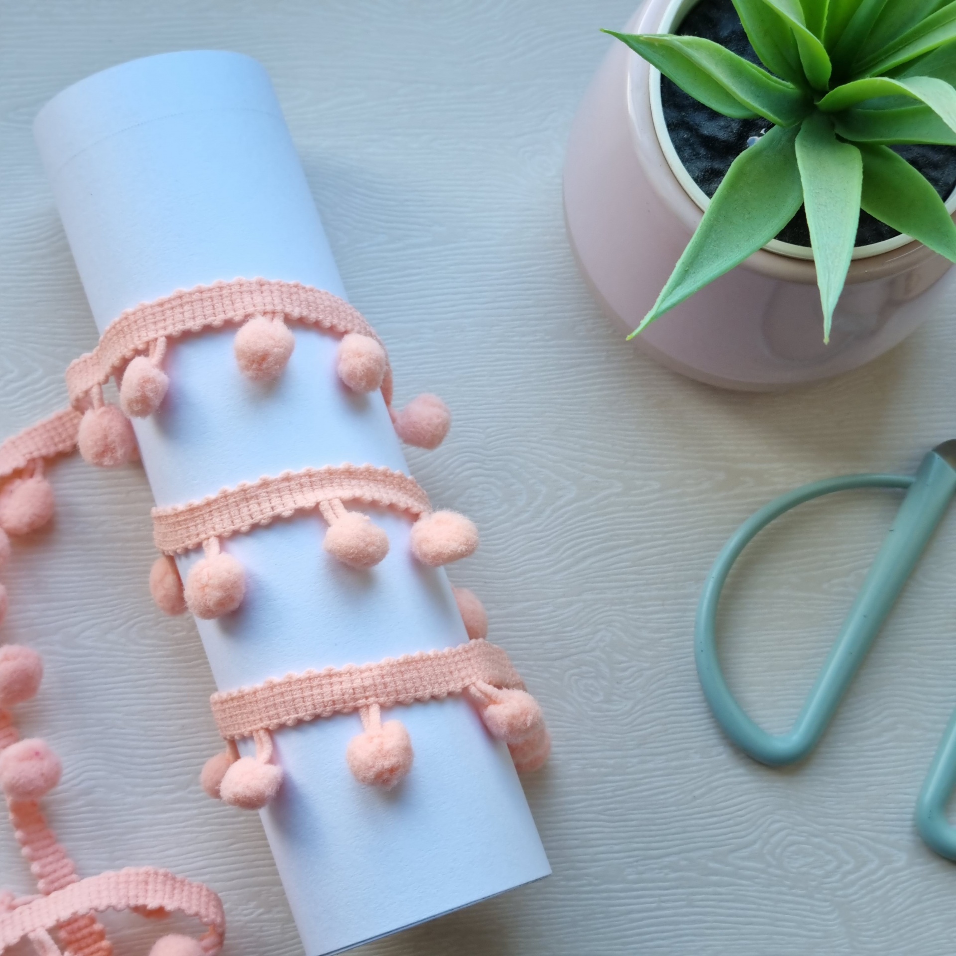 Fita decorativa com pompons, vaso com planta e tesoura sobre superfície de madeira.