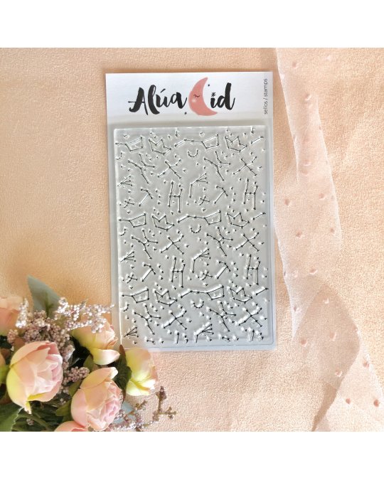 Stencil de constelações em relevo da marca Alua Cid sobre tecido rosa claro próximo a flores.