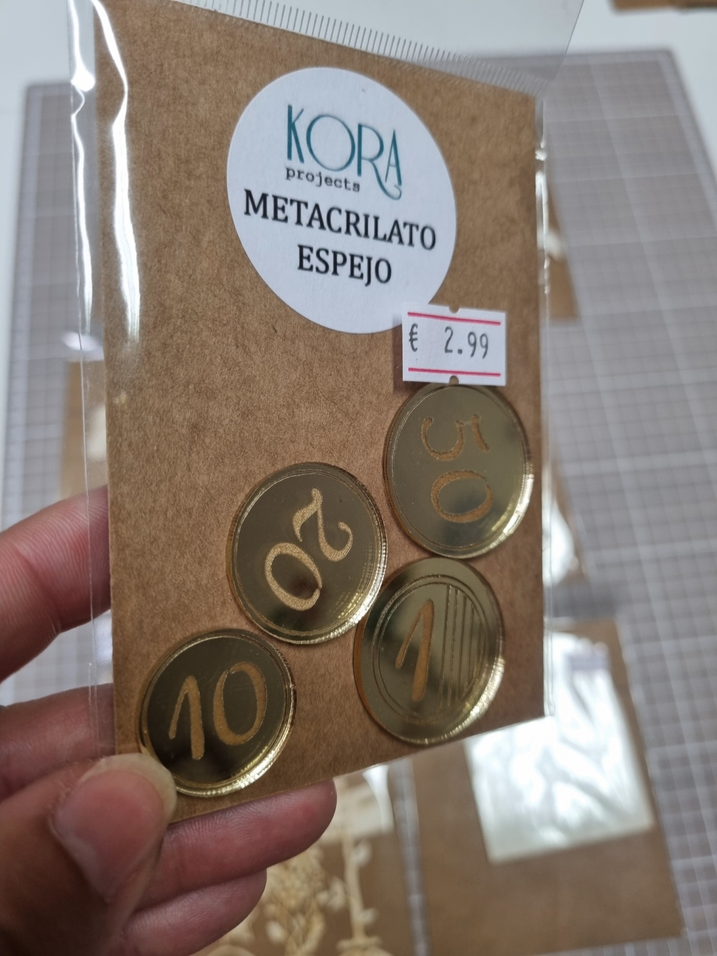 Pacote com moedas douradas de metacrilato espelho e preço em papel castanho com etiqueta redonda.