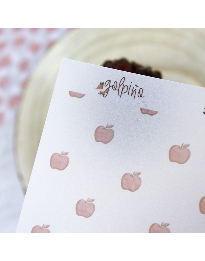 Papel decorado com padrão de maçãs rosas e texto manuscrito 'galpino' sobre fundo de madeira e tecido