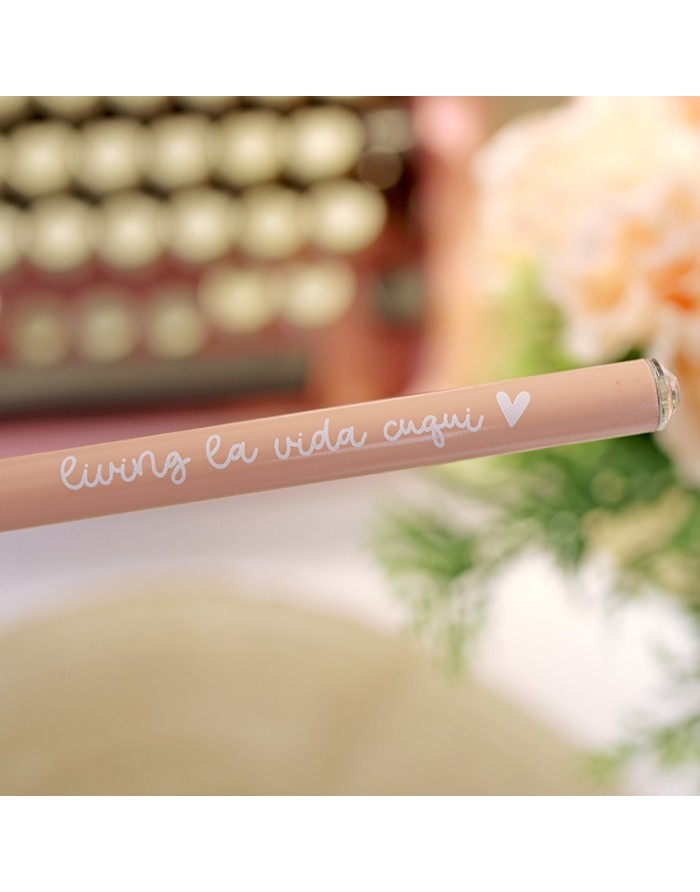 Lápis de cor pêssego com texto 'living la vida cuqui' e coração, fundo desfocado com teclado e flores.