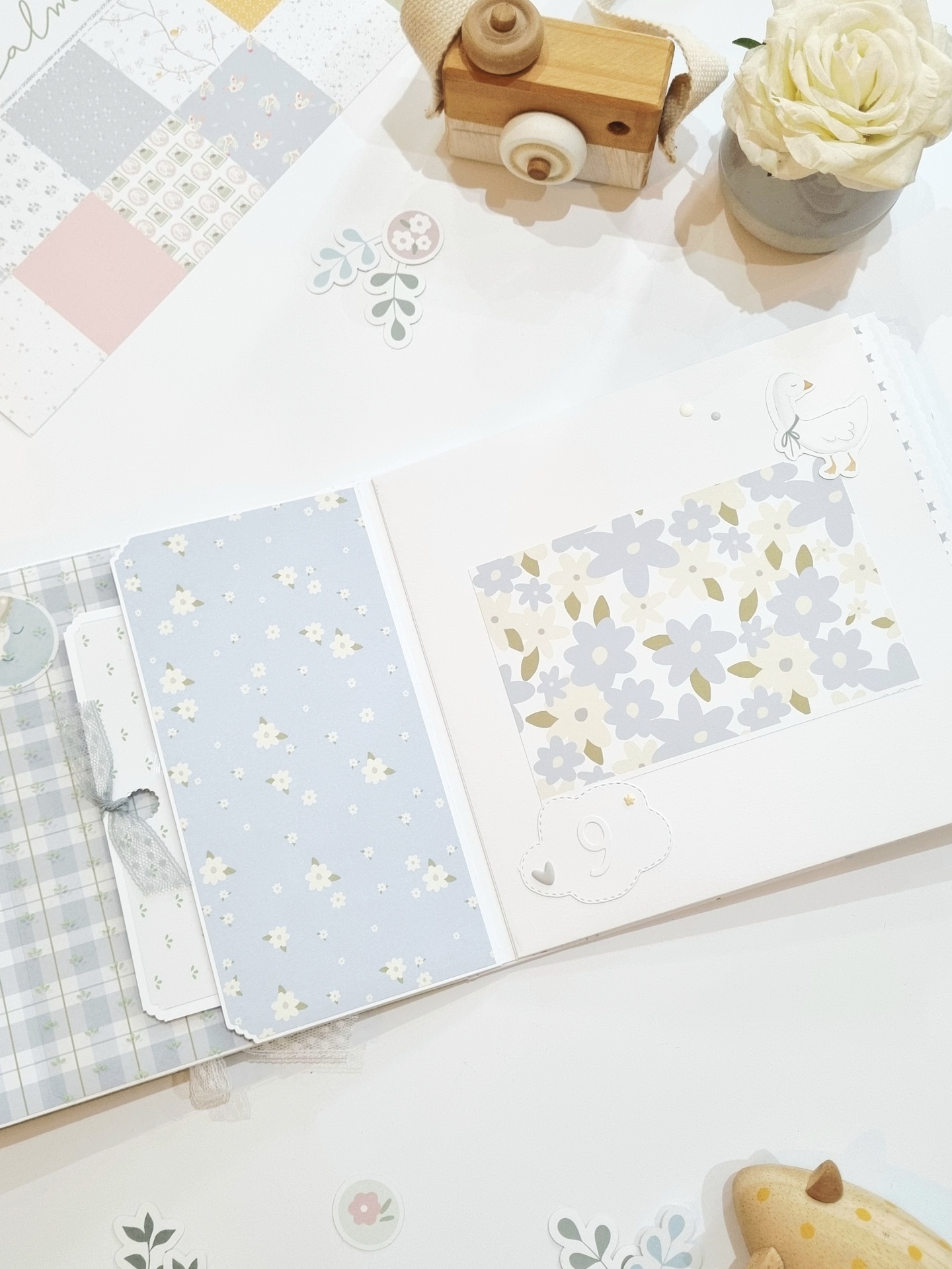 Livro de recordações aberto com decoração floral e adesivos, junto a câmara de madeira e vaso com rosa branca