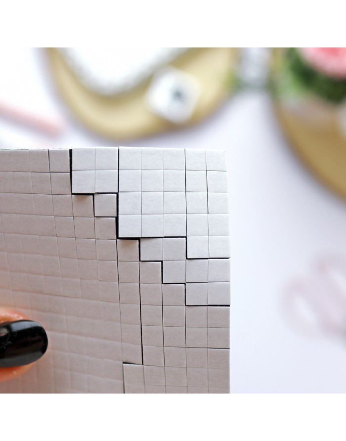 Folha branca quadriculada com recortes em degrau, mão com unhas pretas a segurá-la, fundo desfocado com objetos.