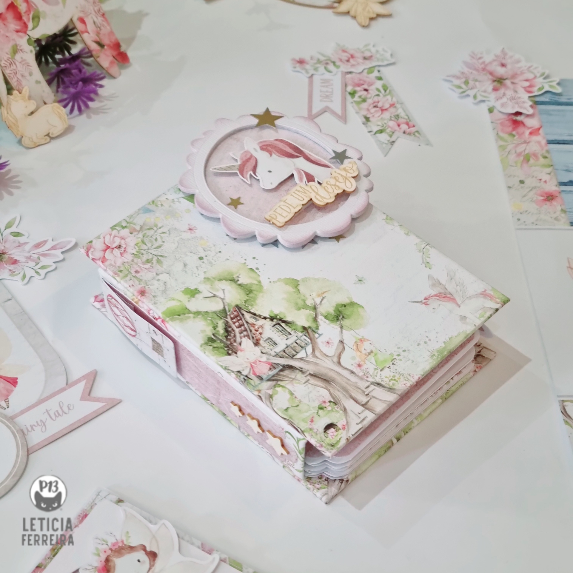 Caderno floral com aplique de unicornio rosa e branco sobre mesa branca com elementos decorativos