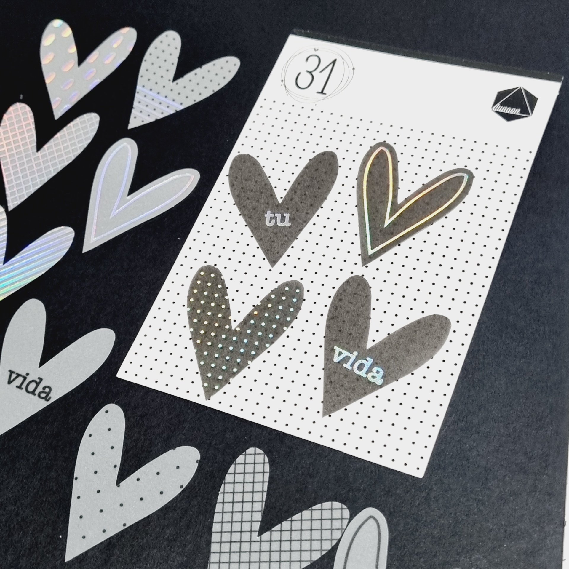 Cartão branco com quatro corações cinza sobre fundo escuro, rodeado por corações recortados em vários padrões e texturas.