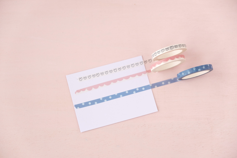 Três rolos de fita adesiva decorativa com corações e um cartão branco sobre superfície rosa