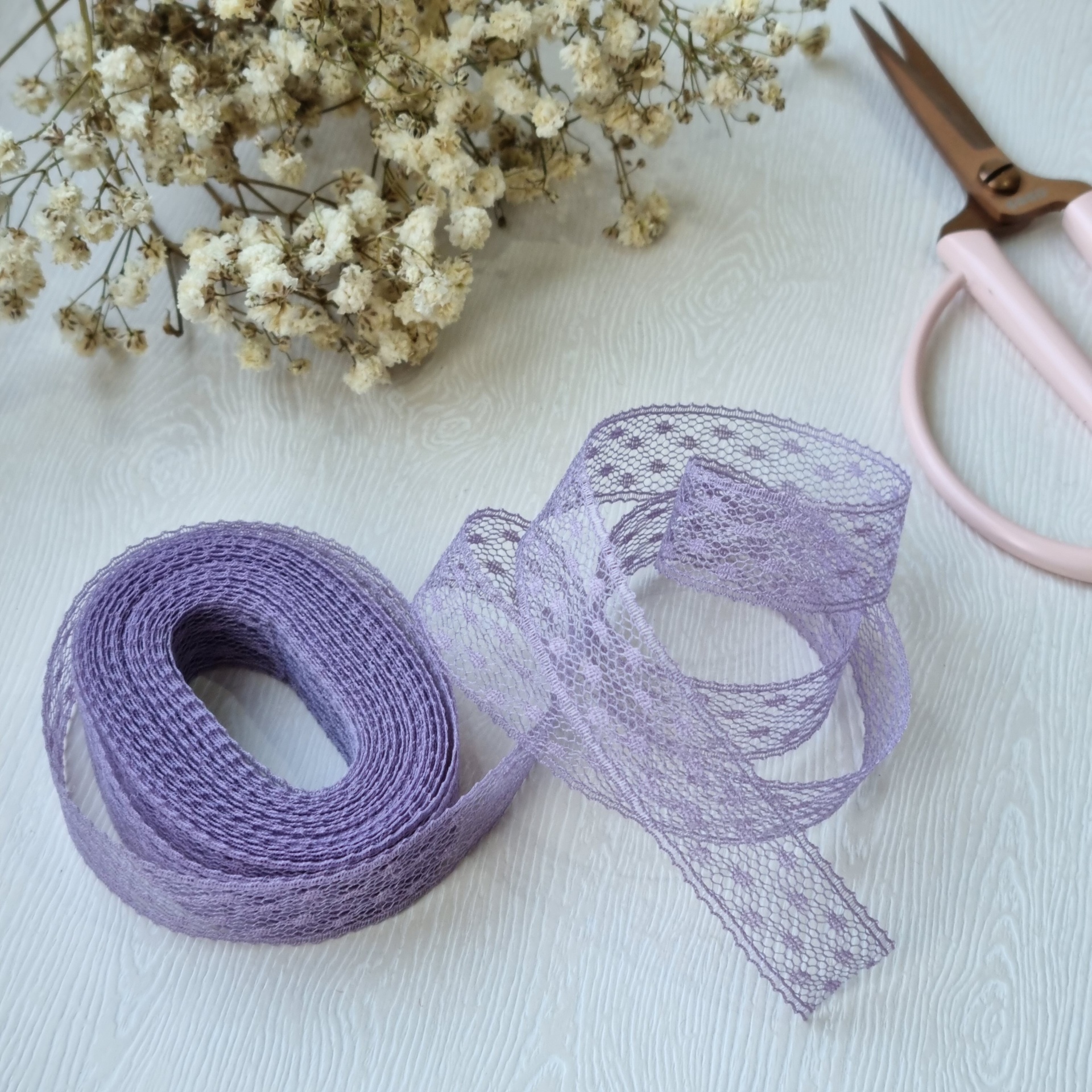Fita de renda roxa desenrolada com flores secas e tesoura em fundo branco