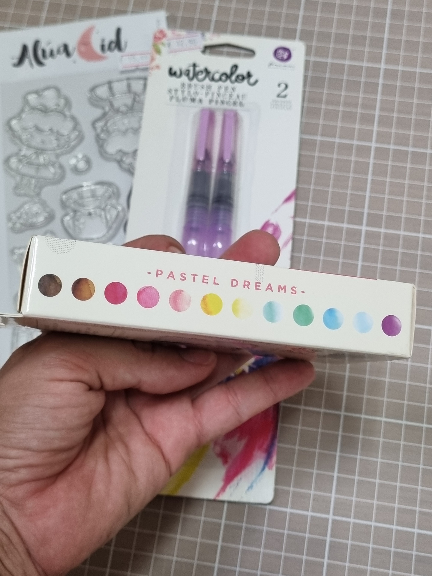 Caixa branca com inscrição PASTEL DREAMS e círculos coloridos em mão, fundo com pincéis de aquarela e carimbos