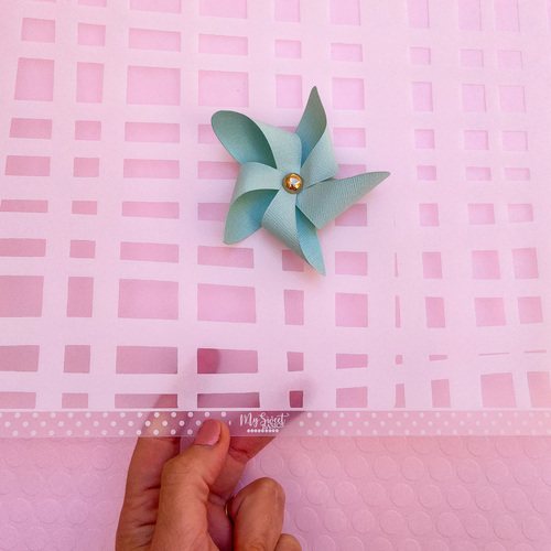 Cata-vento de papel azul claro com joia dourada no centro sobre fundo rosa quadriculado, com mão segurando fita transparente com padrão de bolinhas e texto