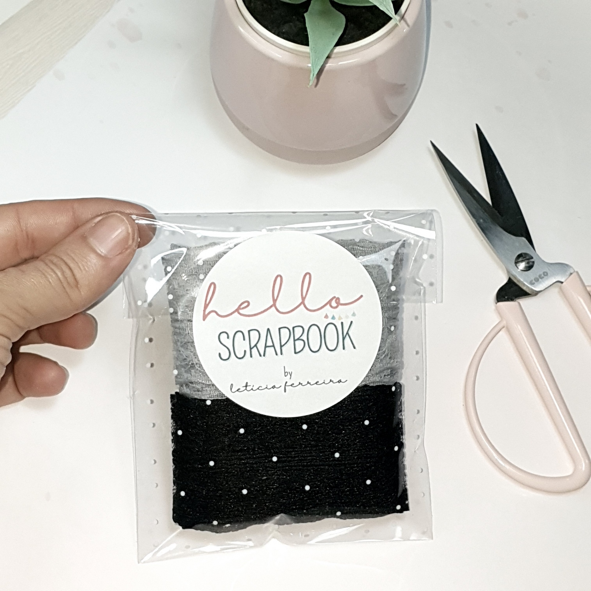 Embalagem transparente com tecido preto e etiqueta branca 'hello SCRAPBOOK', tesoura rosa e vaso com planta