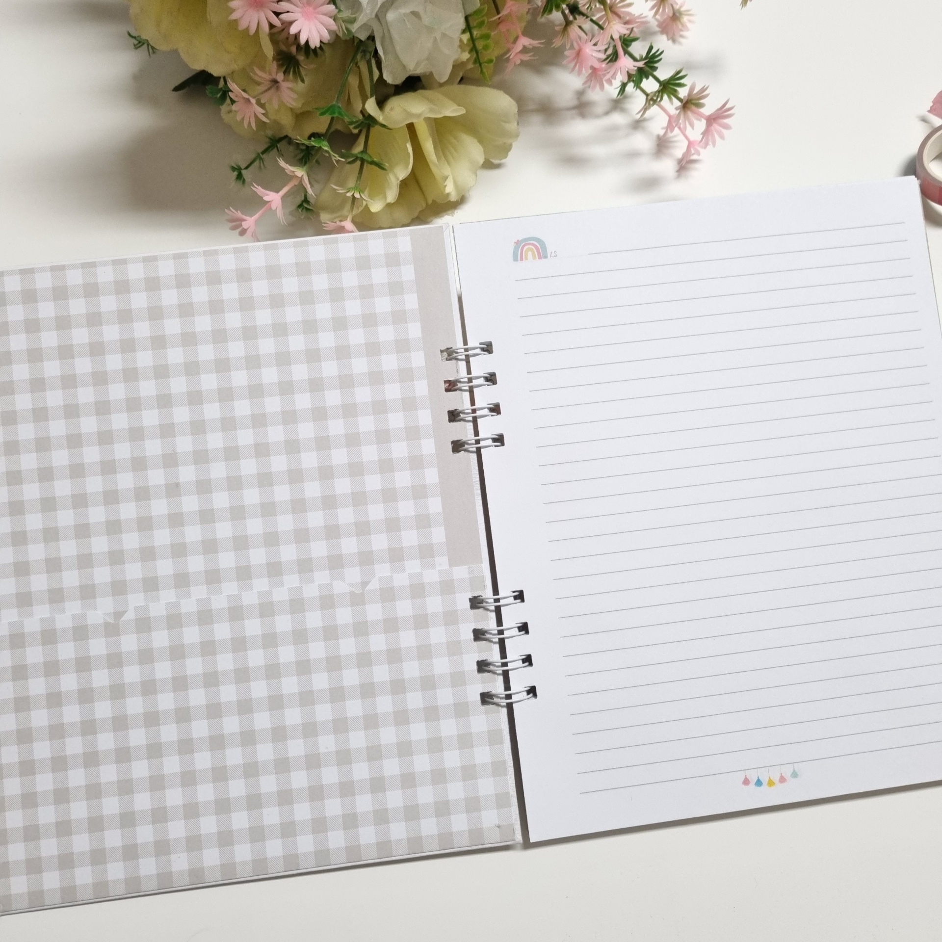 Caderno espiral aberto com páginas pautadas e ilustrações coloridas, capa interna xadrez cinza e branco, flores artificiais ao fundo.