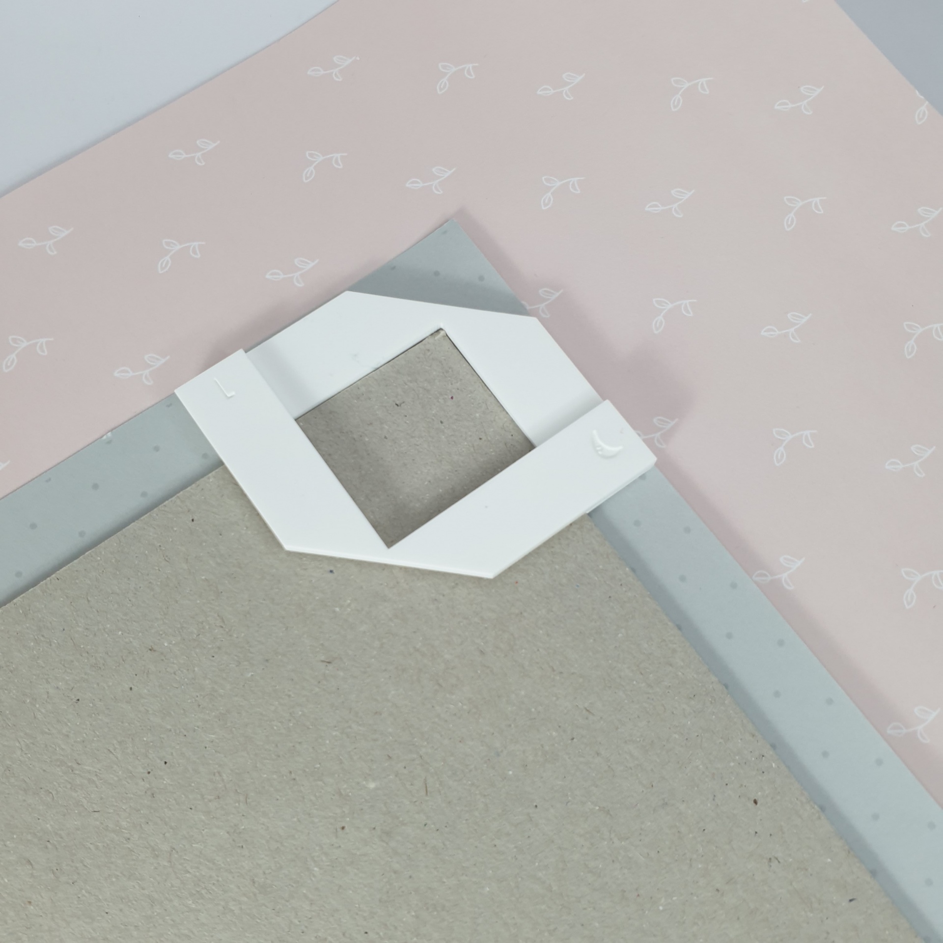 Cantoneira de papelão branca no verso de um quadro com moldura cinza claro sobre fundo rosa com padrões.