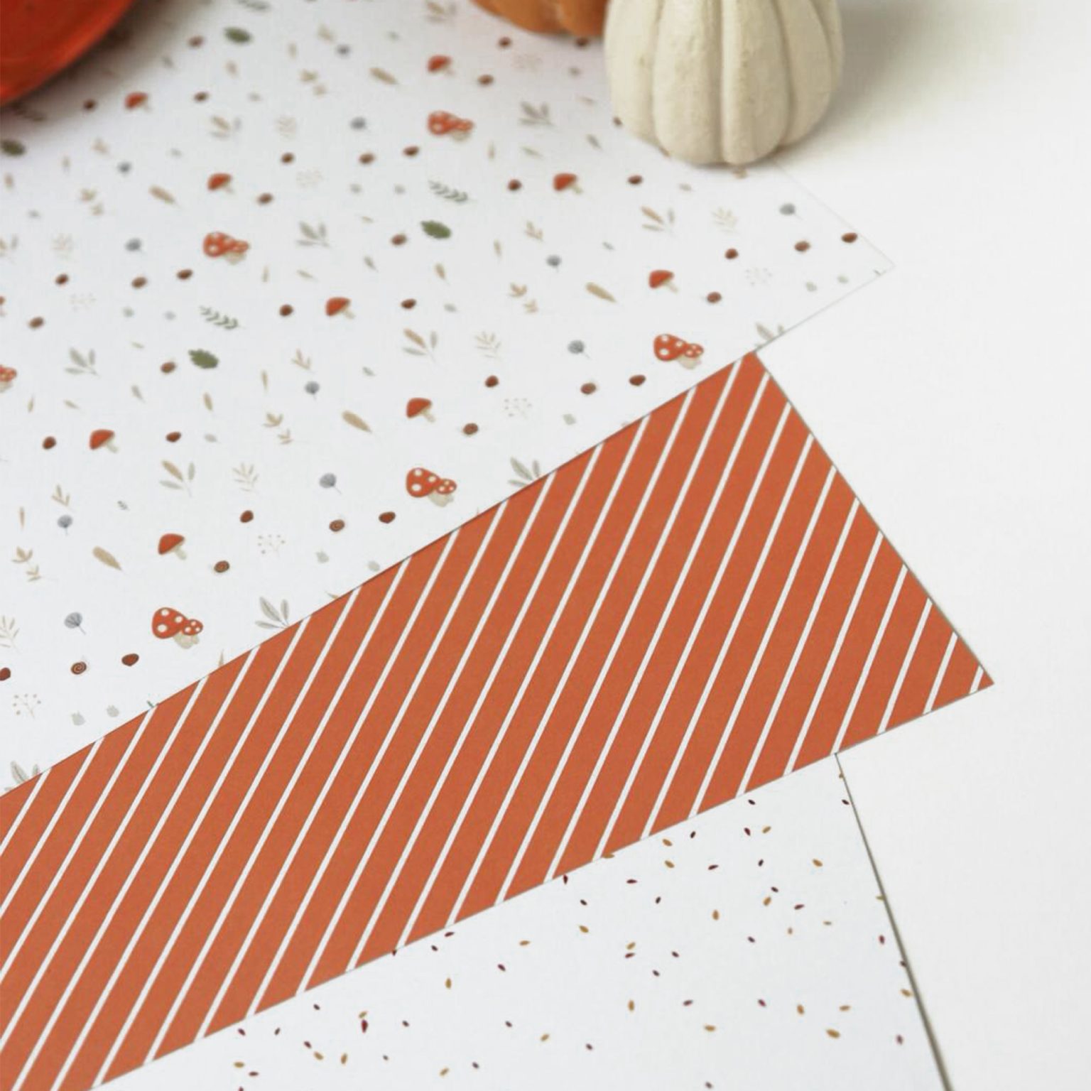 Folhas de papel decorativo com padrões de cogumelos, riscas e manchas, abóbora branca ao lado