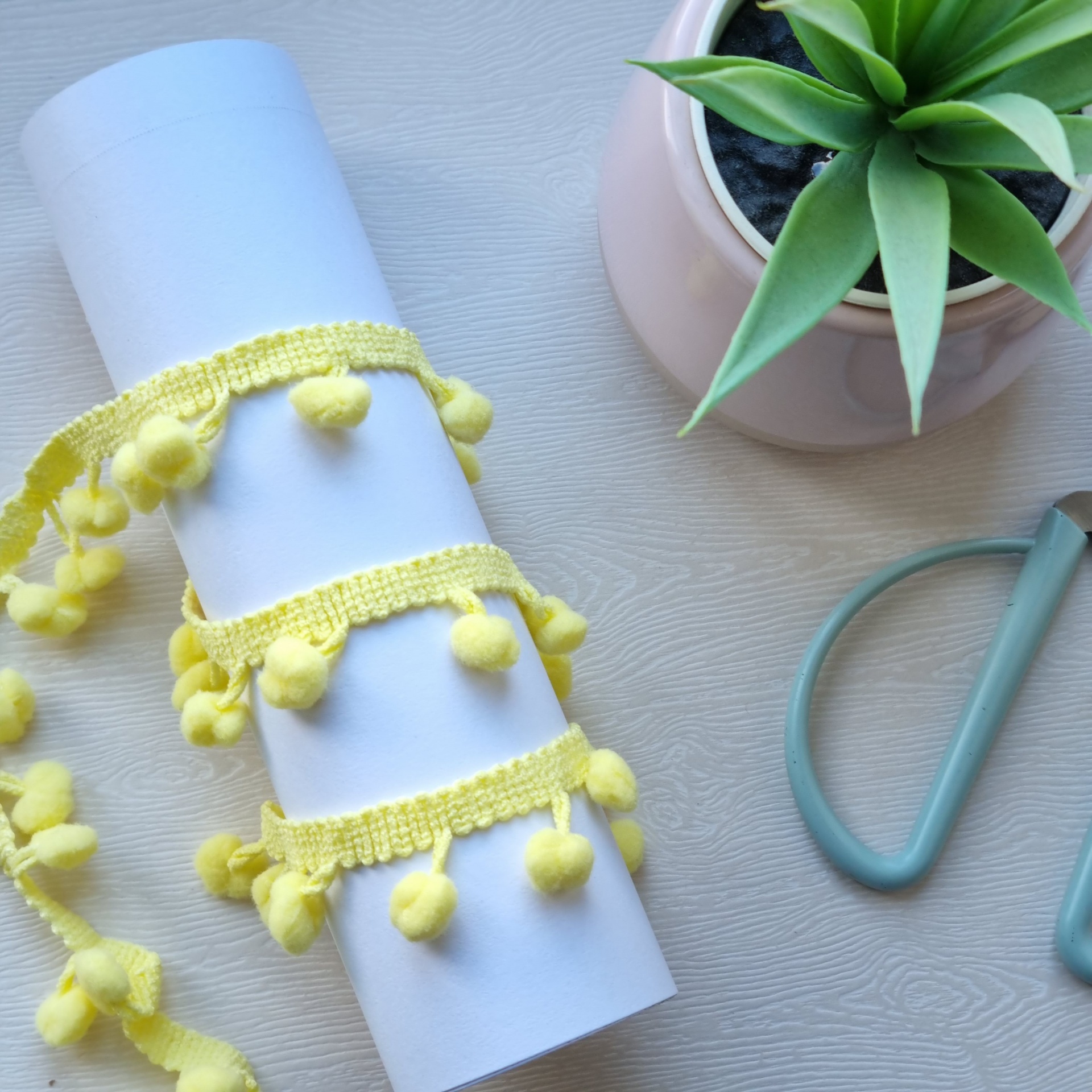 Fita amarela com pompons, rolo de papel branco, planta suculenta em vaso rosa e ferramenta azul sobre superfície clara