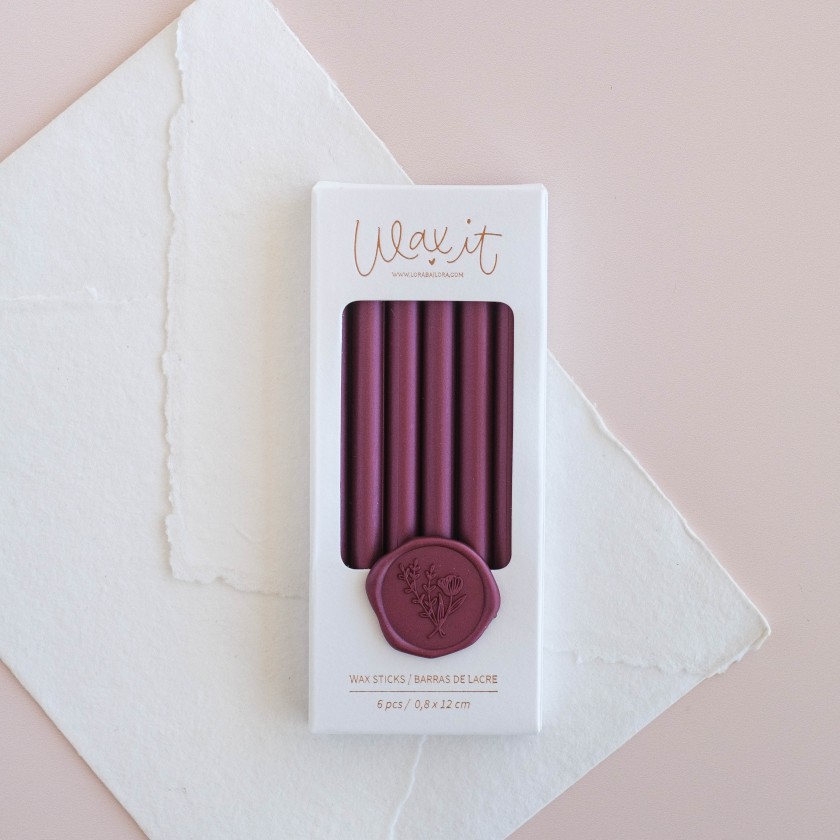 Caixa de varetas de cera cor vinho com selo decorativo em fundo de papel branco e rosa.