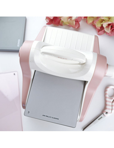 Máquina de corte magnético branca e rosa com unha placa metálica cinza e texto, tesoura rosa listrada e acessórios sobre fundo branco