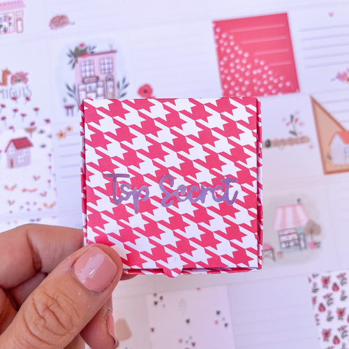 Mão segurando pequena caixa vermelha com padrão pied-de-poule e texto 'top secret' prateado