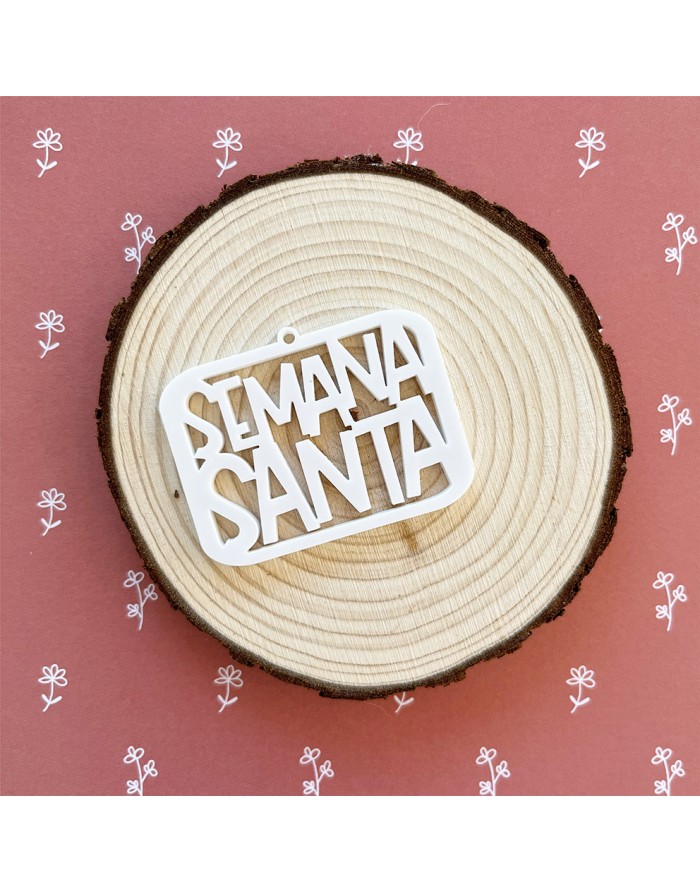 Enfeite branco com 'SEMANA SANTA' sobre uma placa redonda de madeira clara, fundo rosa com flores brancas